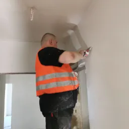 Pracownik w pomarańczowej kamizelce nakłada gładź szpachelką na ścianę w trakcie prac wykończeniowych w jasnym pomieszczeniu.