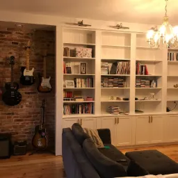 Biała zabudowa meblowa z półkami i szafkami, oświetlona lampami nad półkami, z ceglaną wnęką, w której wiszą gitary elektryczne i akustyczna, oraz stoją wzmacniacze gitarowe, uzupełniona szarą sofą.