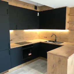 Nowoczesna kuchnia z czarnymi frontami szafek i blatem imitującym drewno, oświetlenie LED pod szafkami wiszącymi, czarna bateria i zlew, płyta indukcyjna i piekarnik w zabudowie, wisząca żarówka...