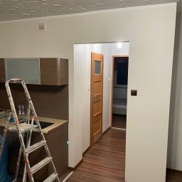 Filip Tuszyński FIL-BUD Usługi Ogólnobudowlane - Wnętrze mieszkania w trakcie odświeżania ścian, widoczna drabina malarska przy zabudowie kuchennej, świeżo pomalowana ściana w kolorze złamanej bieli, wzorzysty sufit, drewniana podłoga i drzwi...