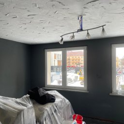 Filip Tuszyński FIL-BUD Usługi Ogólnobudowlane - Pokój w trakcie prac malarskich: ściany pomalowane na ciemny kolor, sufit z dekoracyjną fakturą, meble zabezpieczone folią, widoczne narzędzia malarskie i puszki z farbą.