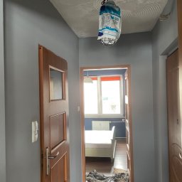 Filip Tuszyński FIL-BUD Usługi Ogólnobudowlane - Przygotowanie do malowania: Pokój z szarymi ścianami, zabezpieczoną folią podłogą, zdjętą lampą, odsłoniętą żarówką i wiszącą reklamą taśmy malarskiej.