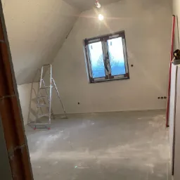 Poddasze w trakcie prac wykończeniowych: ściany i skosy obłożone płytami kartonowo-gipsowymi, widoczna drabina, okno z widokiem na drzewa oraz wystające przewody elektryczne.