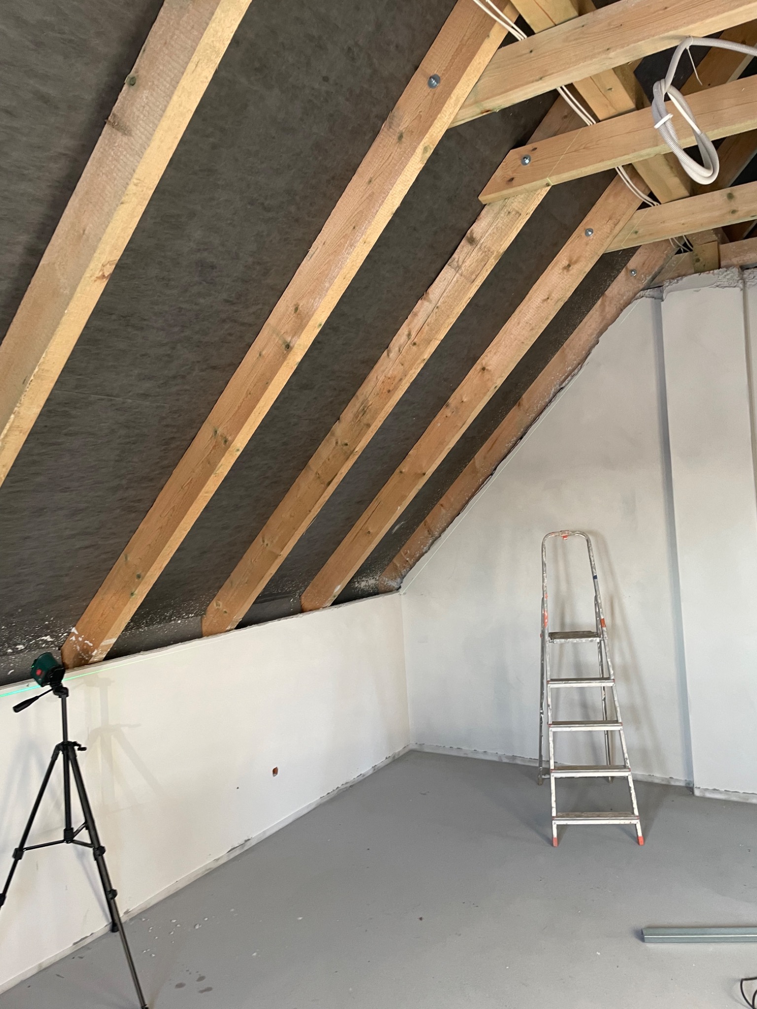 Poddasze w trakcie prac wykończeniowych: widoczne drewniane krokwie z izolacją, gładkie, białe ściany, poziomica laserowa na statywie i aluminiowa drabina. Szare wykończenie podłogi.