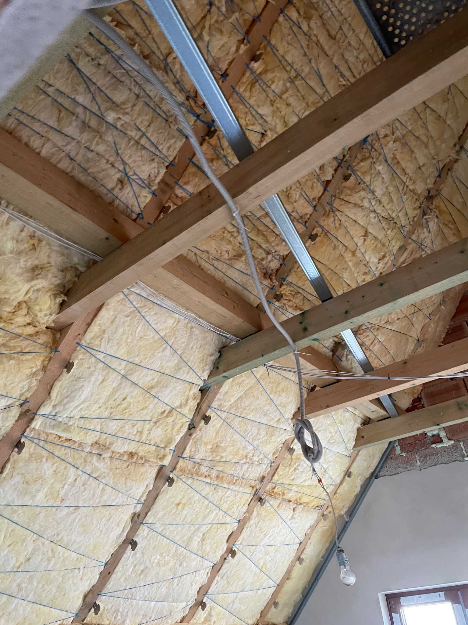 Izolacja poddasza wełną mineralną, zabezpieczona sznurkiem, widoczne drewniane krokwie i metalowe profile, surowy stan wykończenia, wisząca żarówka.