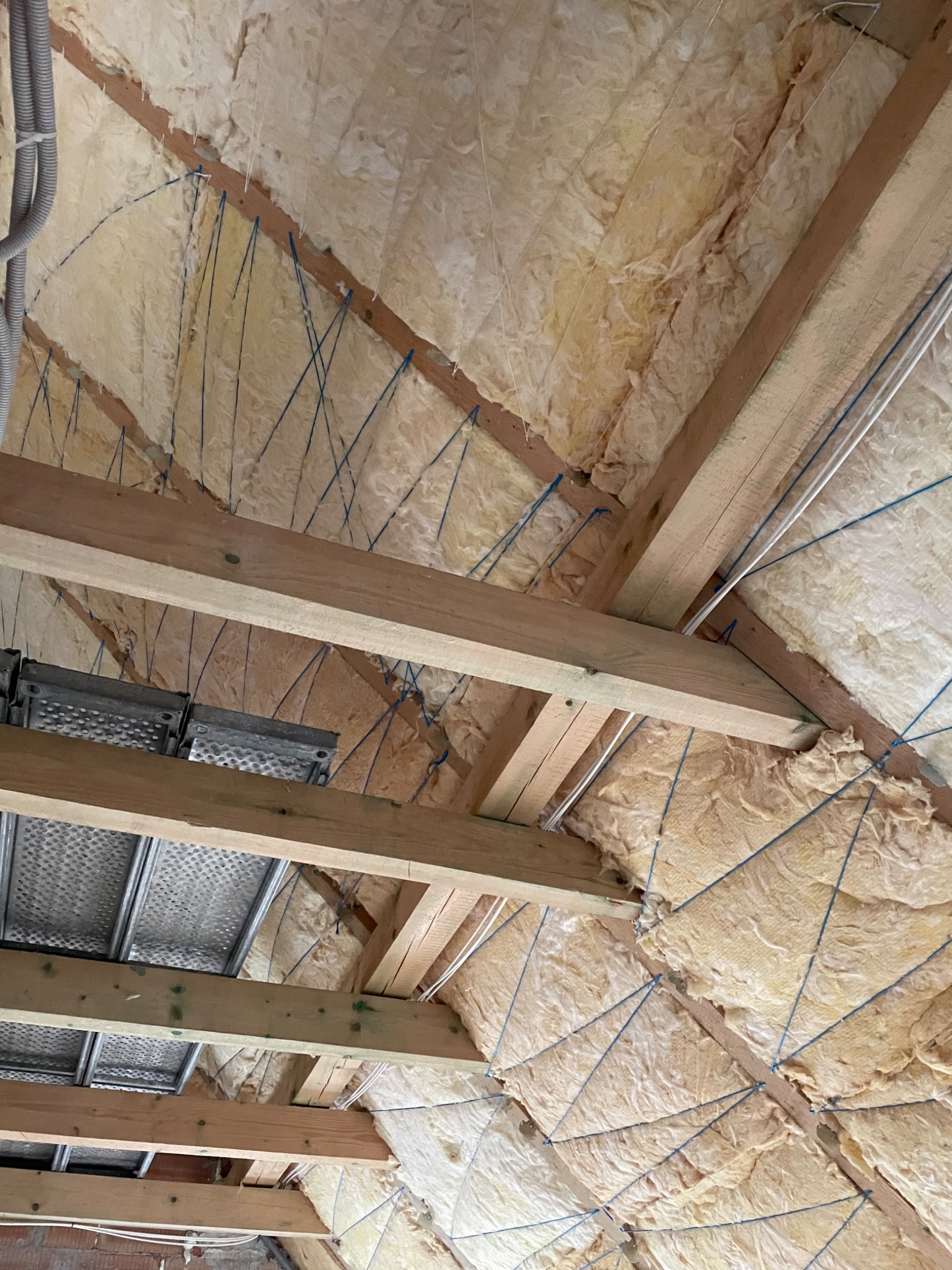 Izolacja poddasza wełną mineralną mocowaną sznurkami do drewnianej konstrukcji, widoczny fragment rusztowania.