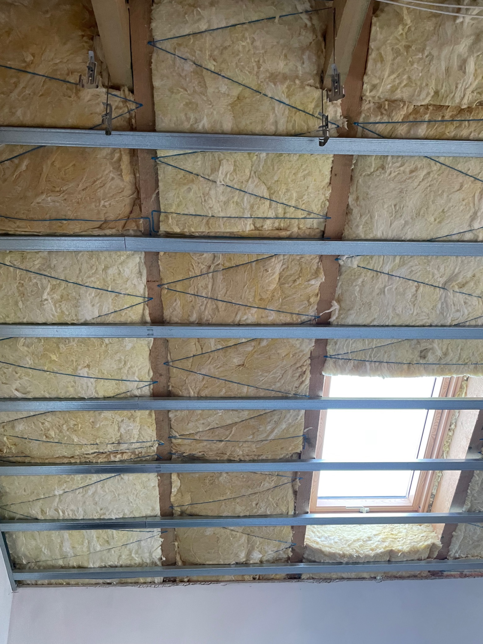 Izolacja poddasza wełną mineralną, widoczne metalowe profile, drewniane belki i okno dachowe z otwartym skrzydłem, niebieski sznurek podtrzymujący izolację.