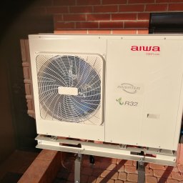 Synterm Artur Broniś - Zewnętrzna jednostka pompy ciepła Aiwa zamontowana na wspornikach przy ceglanym budynku, widoczny wentylator z niebieskimi elementami, logo i oznaczenie R32.