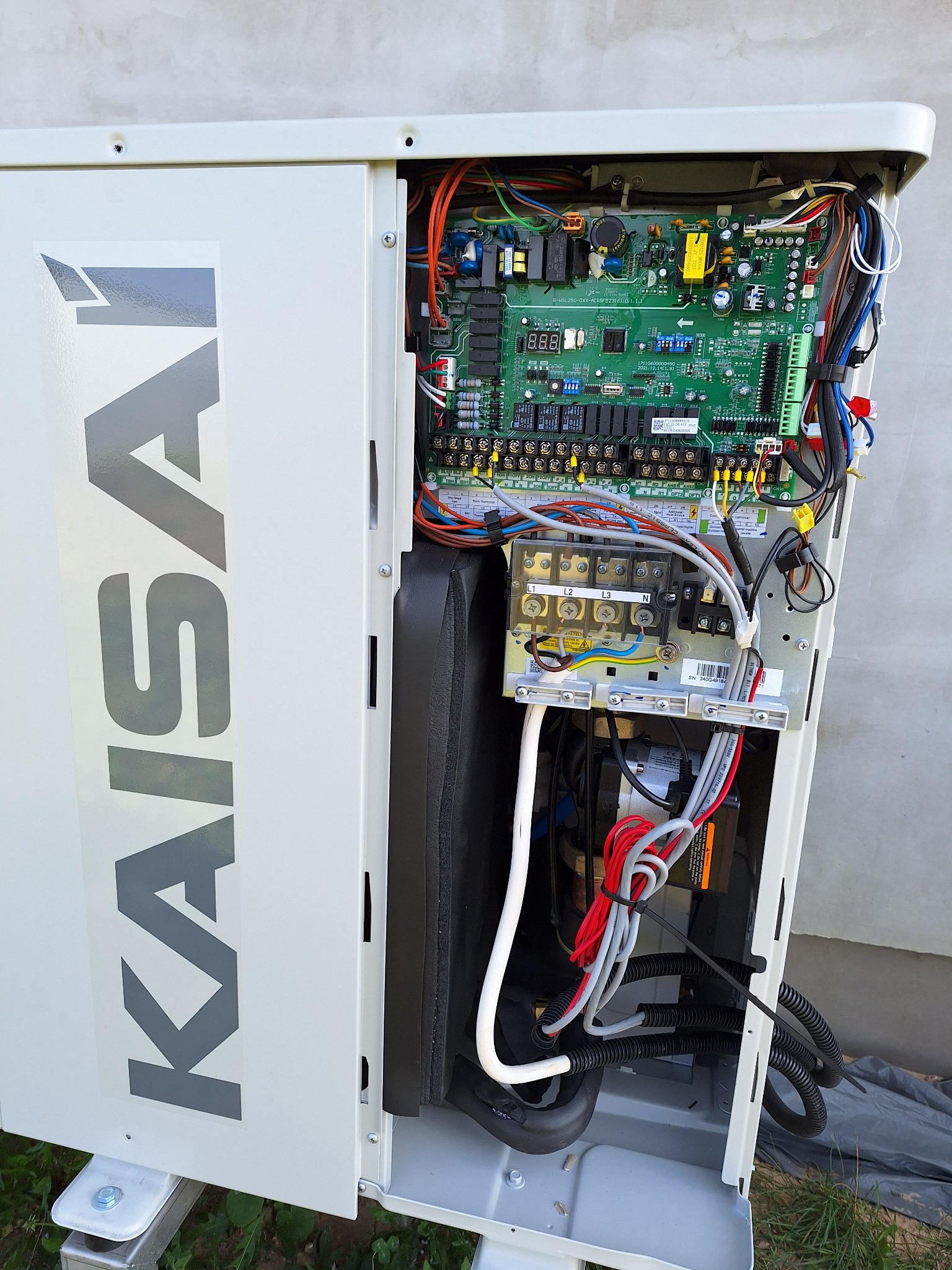 Otwarta obudowa jednostki zewnętrznej z widocznymi elementami elektronicznymi, przewodami i tabliczką znamionową Kaisai, zamontowana na zewnątrz budynku.
