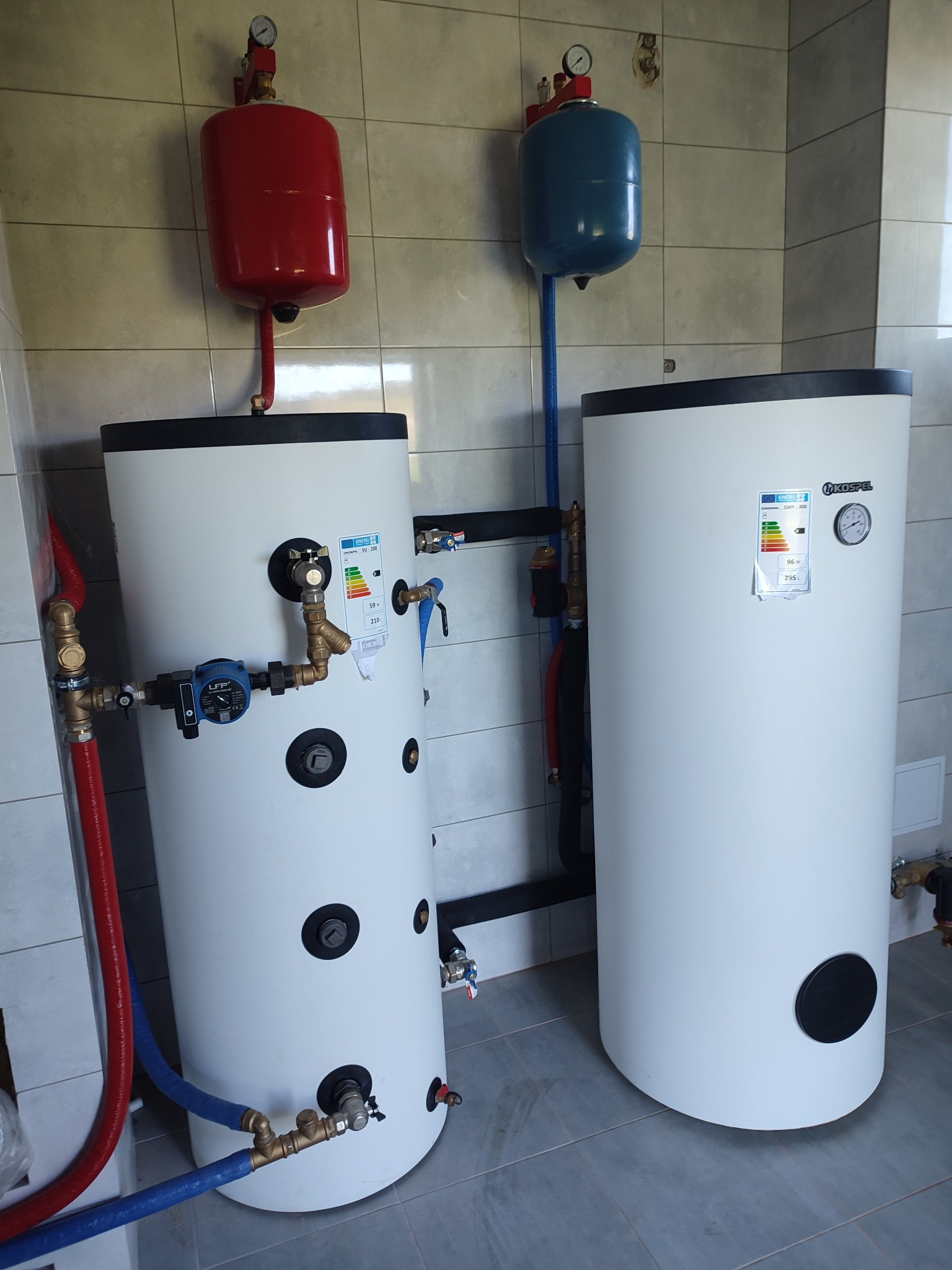 Dwa białe zasobniki ciepłej wody użytkowej z podłączonymi rurami (czerwona i niebieska), manometry na zbiornikach wyrównawczych, płytki ceramiczne na ścianie w tle.