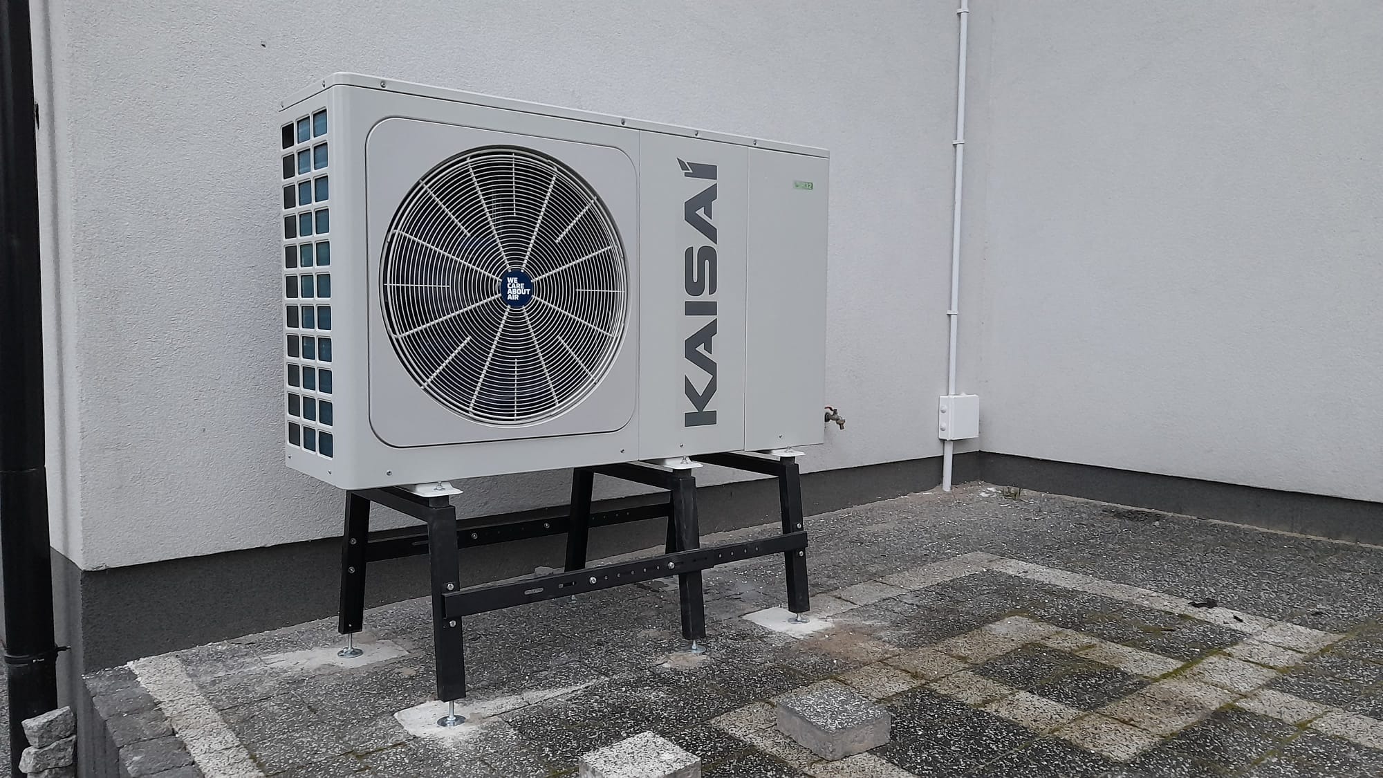 Szara pompa ciepła marki Kaisai zamontowana na czarnej, metalowej konstrukcji na szarym brukowanym podwórku przy białej ścianie budynku.