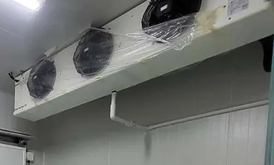 Biała, podwieszana jednostka chłodnicza z trzema wentylatorami, częściowo owinięta folią ochronną, zamontowana w chłodni lub magazynie spożywczym o ścianach z paneli warstwowych.