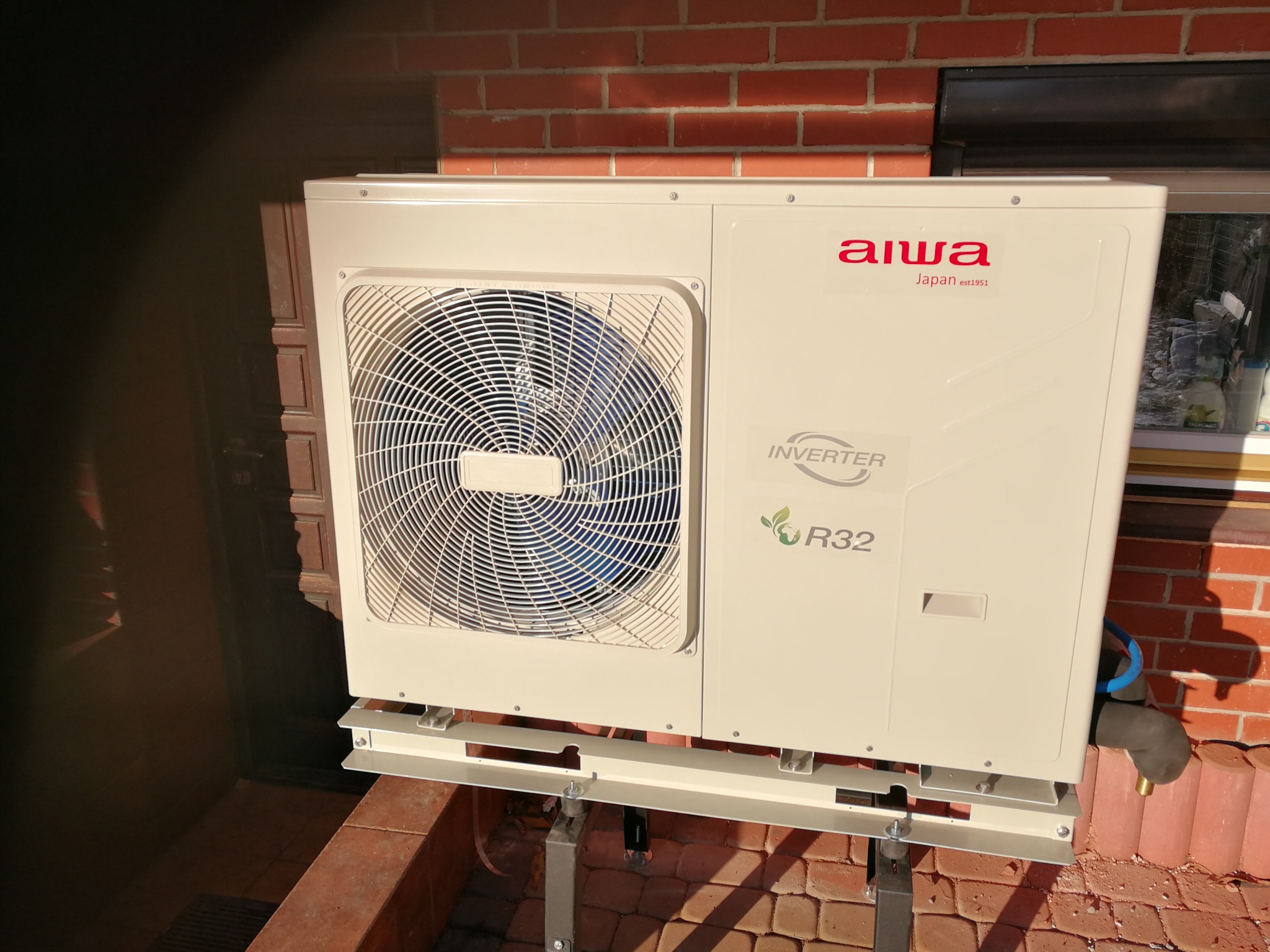 Zewnętrzna jednostka pompy ciepła Aiwa zamontowana na wspornikach przy ceglanym budynku, widoczny wentylator z niebieskimi elementami, logo i oznaczenie R32.
