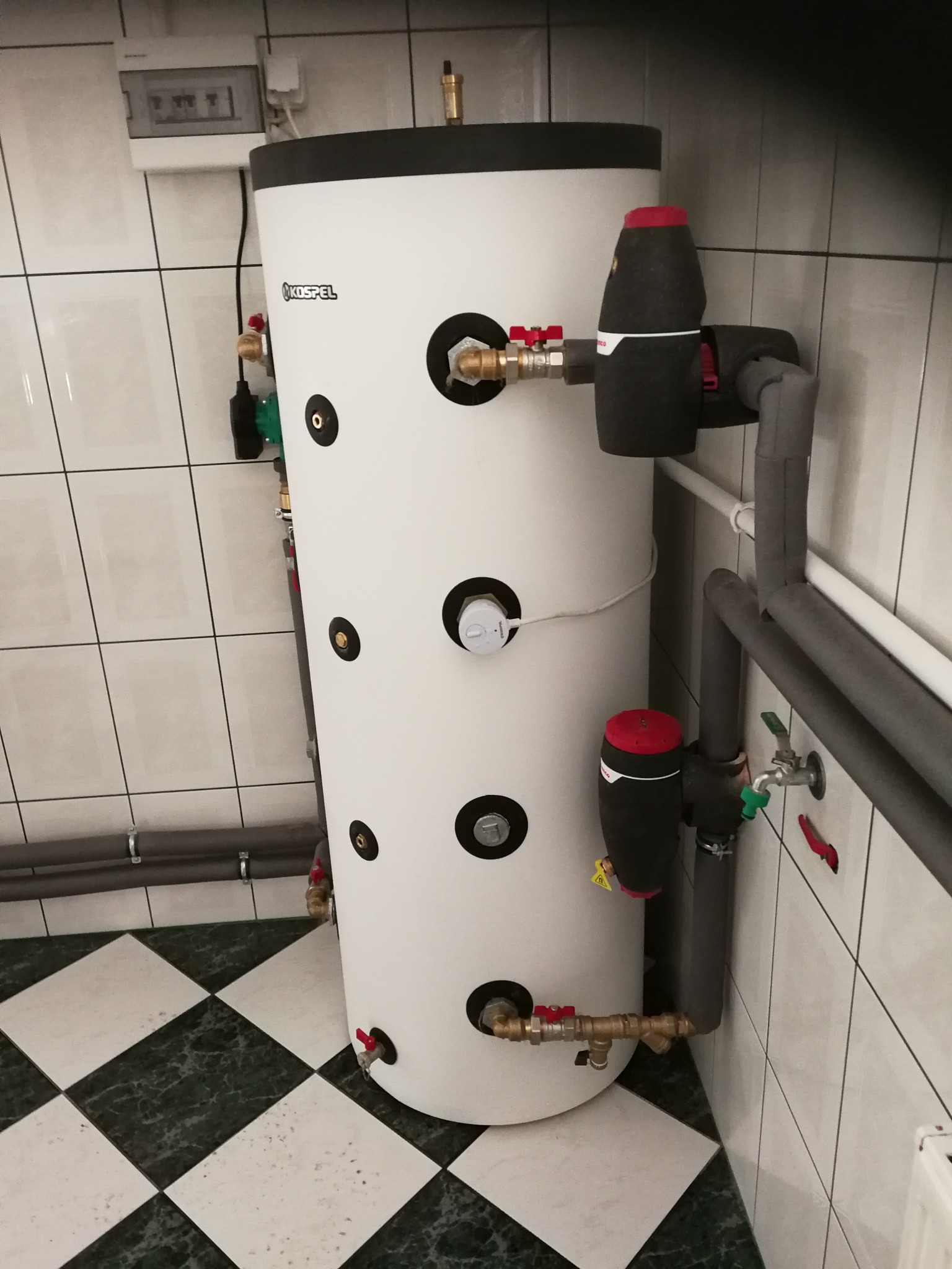 Biały, cylindryczny zasobnik ciepłej wody użytkowej Kospel z czarnymi okrągłymi zaślepkami i przyłączonymi rurami, zaworami i pompami obiegowymi w kotłowni z biało-zieloną szachownicą na podłodze...