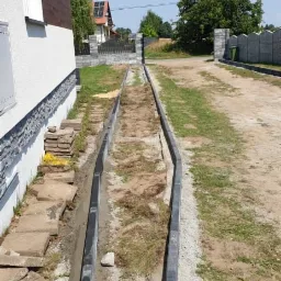 Montaż obrzeży betonowych wzdłuż podjazdu, widoczny fragment budynku z szarą elewacją i ułożone kostki brukowe w tle.