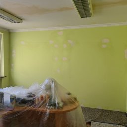 Darlbud Mateusz Dul - Pokój z zielonymi ścianami w trakcie przygotowania do malowania: ściany z zagruntowanymi miejscami po szpachlowaniu, stół przykryty folią ochronną, widoczne ślady po zalaniu na suficie.