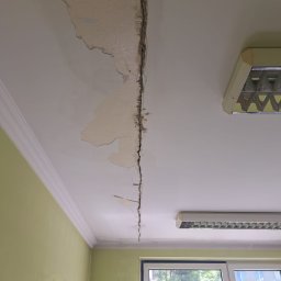 Darlbud Mateusz Dul - Pęknięcie i odpadający tynk na suficie w pomieszczeniu z oknem i lampą fluorescencyjną, widoczne uszkodzenia wymagające naprawy.