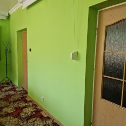 Darlbud Mateusz Dul - Ściana pomalowana na jasnozielono z drewnianą stolarką drzwiową i wzorzystą wykładziną podłogową. Widoczny włącznik światła z łańcuszkiem, gniazdko elektryczne i wieszak na ubrania.