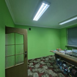 Darlbud Mateusz Dul - Pomalowane na jaskrawo zielono wnętrze pomieszczenia z otwartymi drewnianymi drzwiami z szybą ornamentową, widoczne narzędzia i materiały pod stołem, wzorzysta podłoga.