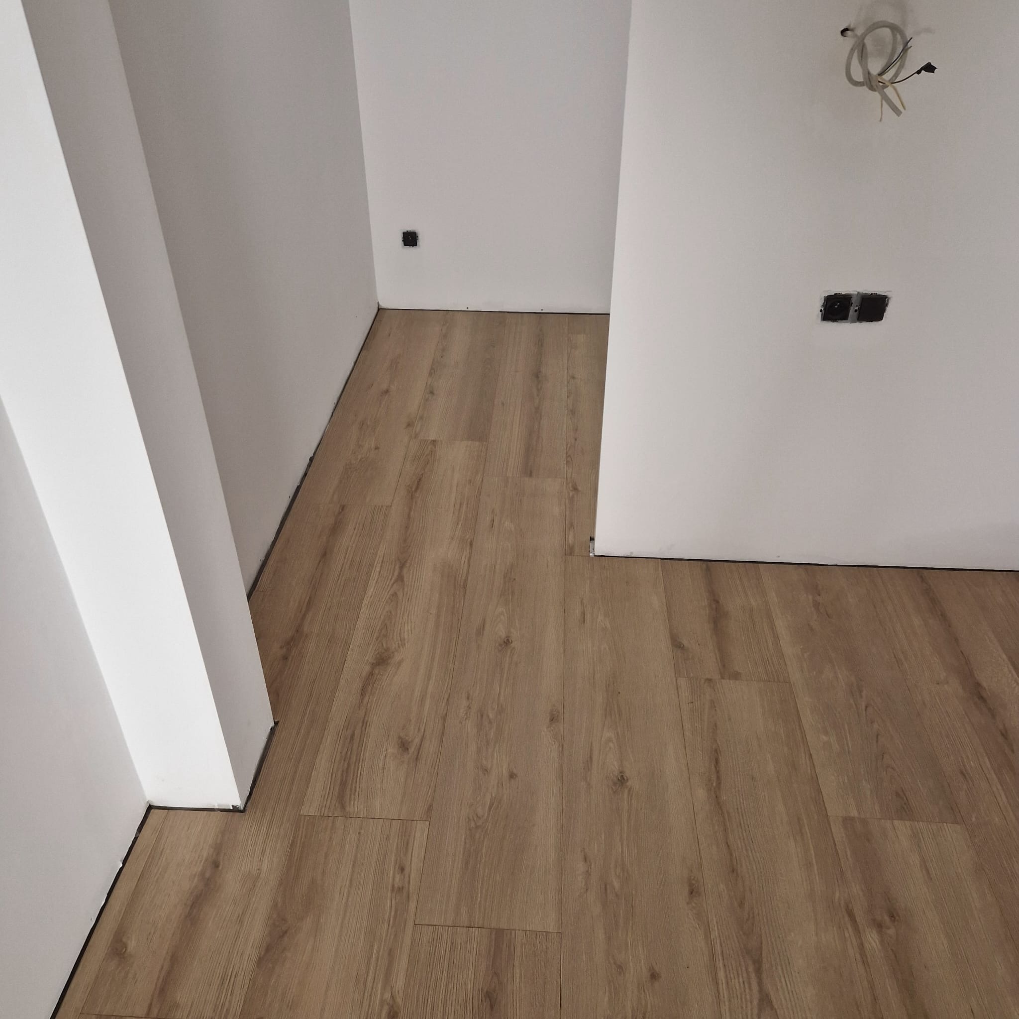Ułożona podłoga z paneli imitujących drewno w pomieszczeniu z białymi ścianami i widocznymi gniazdkami elektrycznymi oraz wystającymi przewodami ze ściany.