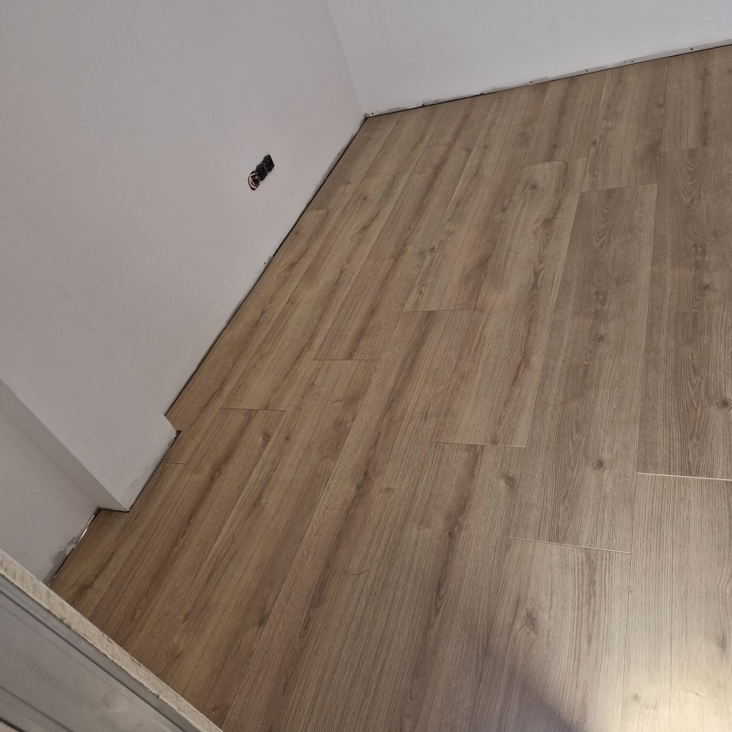 Ułożona podłoga z paneli imitujących drewno w narożniku pokoju, widoczne niedociągnięcia przy ścianie i gniazdko elektryczne bez osłonki.