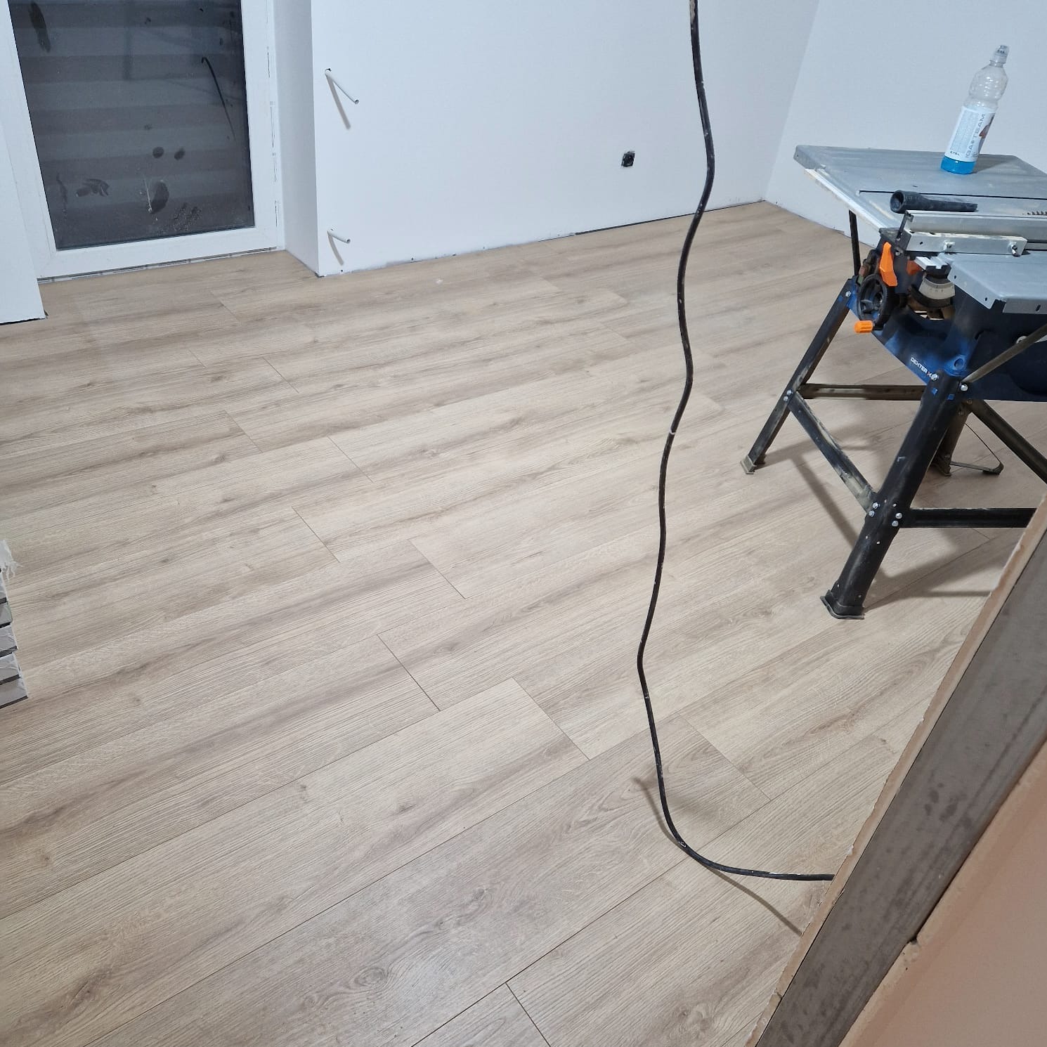 Jasne panele podłogowe ułożone w pokoju, widoczna piła stołowa na stojaku i kabel zasilający, ściany białe z widocznymi śladami po wierceniu.