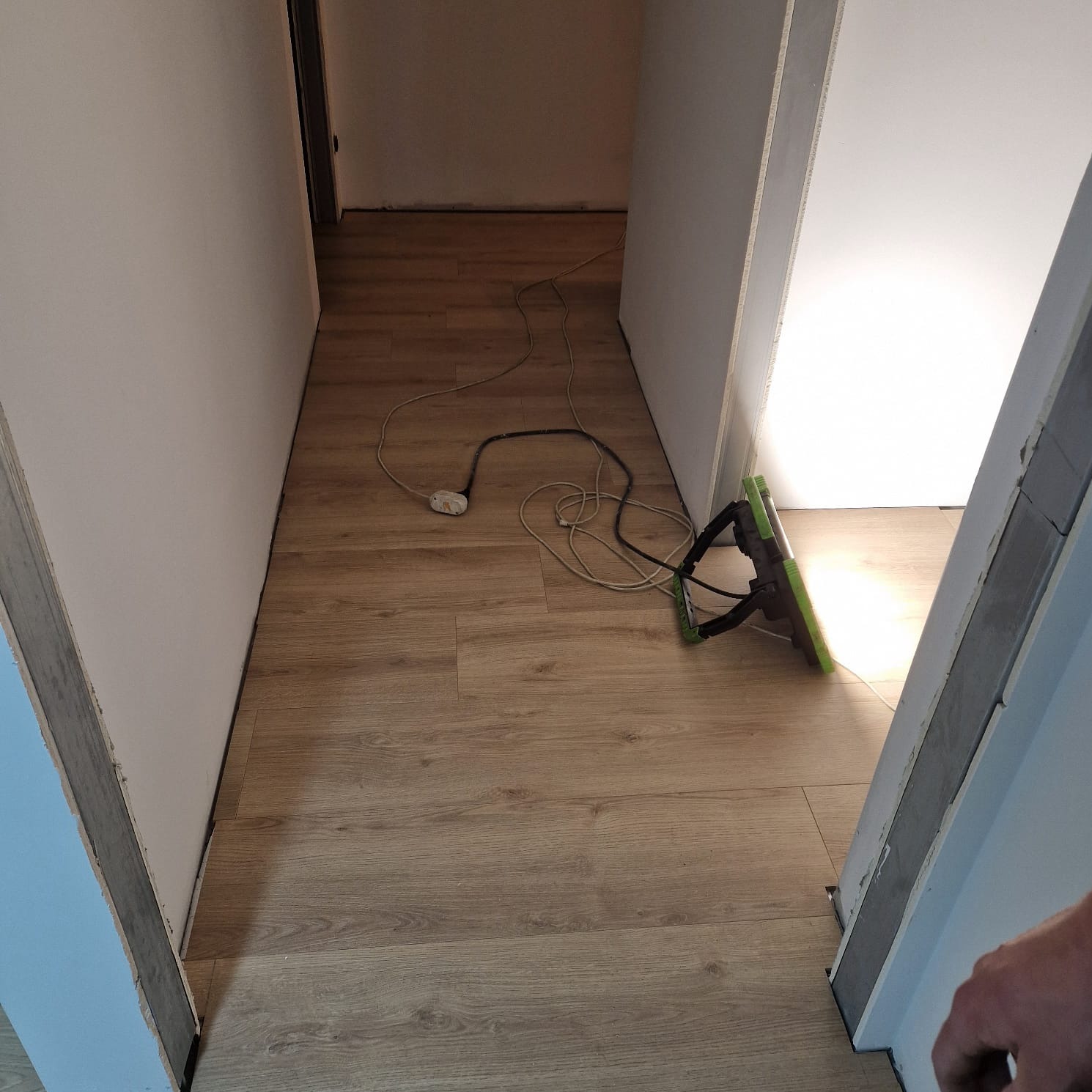 Korytarz w trakcie prac wykończeniowych z widoczną lampą roboczą, kablami na podłodze z paneli i przygotowanymi ścianami pod malowanie.