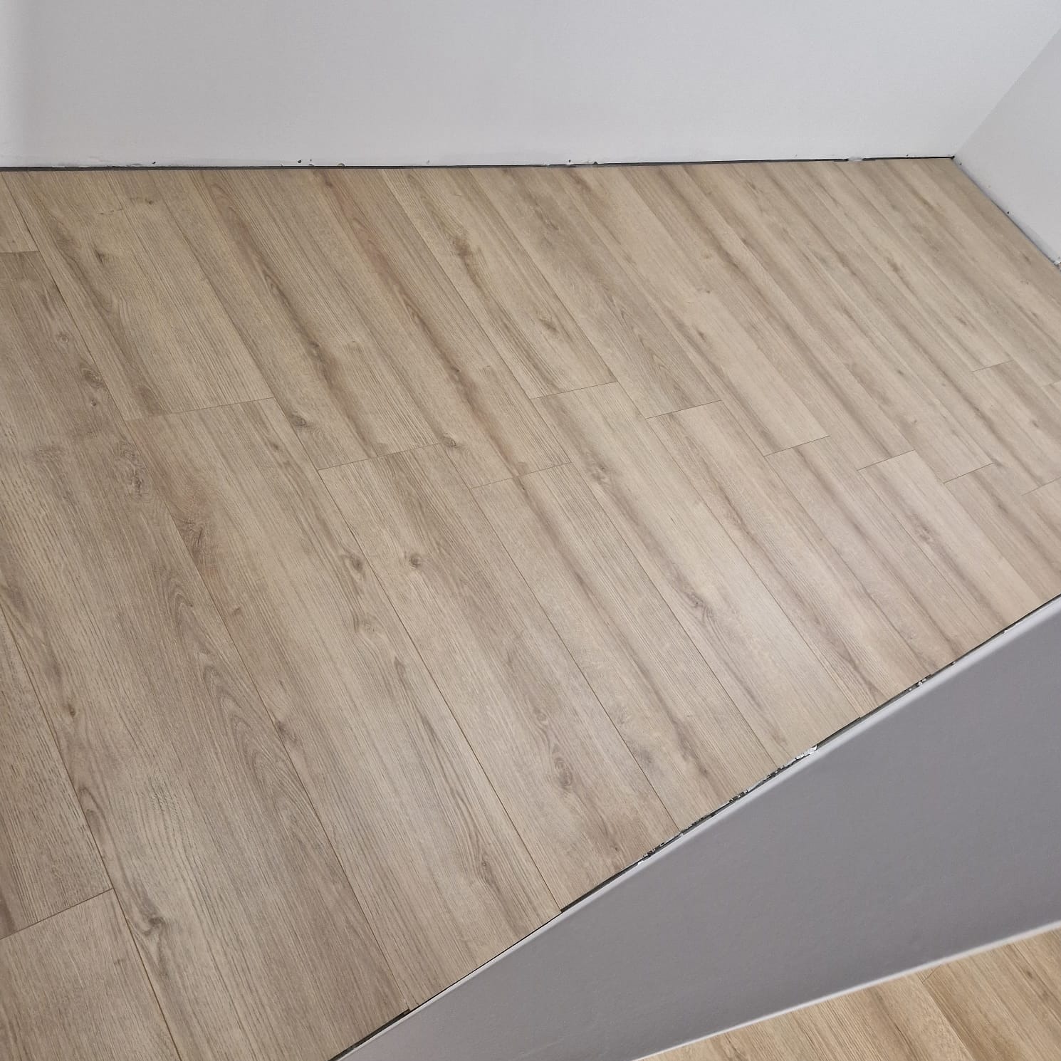 Ukończona podłoga z paneli laminowanych w odcieniach jasnego drewna, widok z góry, ściana pomalowana na biało, listwa przypodłogowa szara.