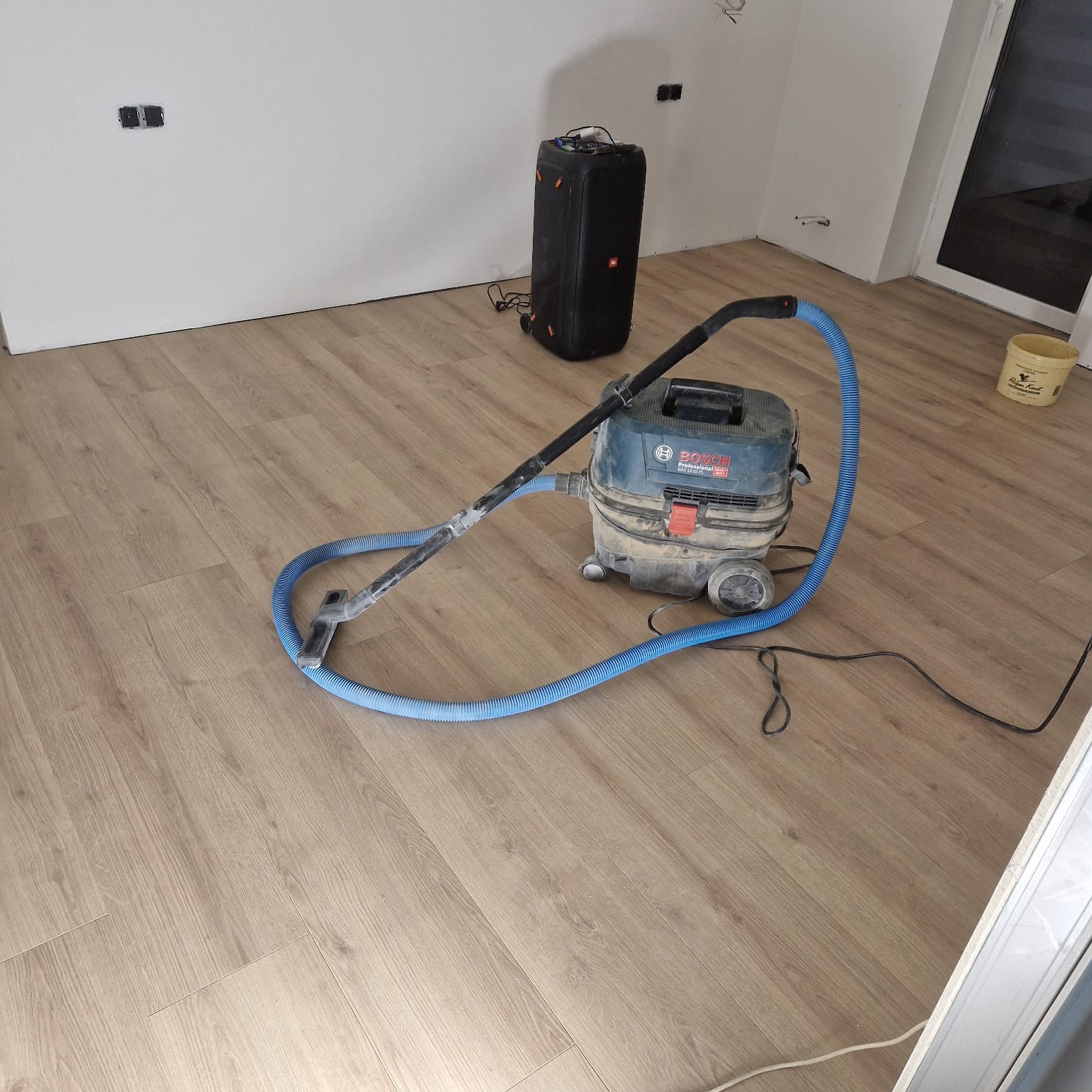 Podłoga z paneli po remoncie, czyszczona przemysłowym odkurzaczem Bosch z niebieskim wężem, w tle białe ściany z gniazdkami i głośnik JBL.