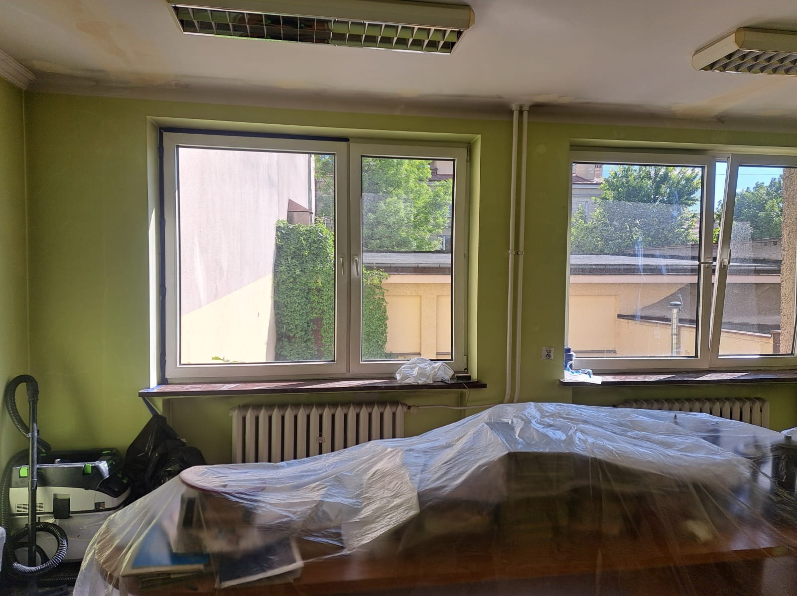 Pokój przed remontem: okna z widokiem na zieleń, grzejniki, podłoga i meble przykryte folią ochronną, widoczny odkurzacz przemysłowy.