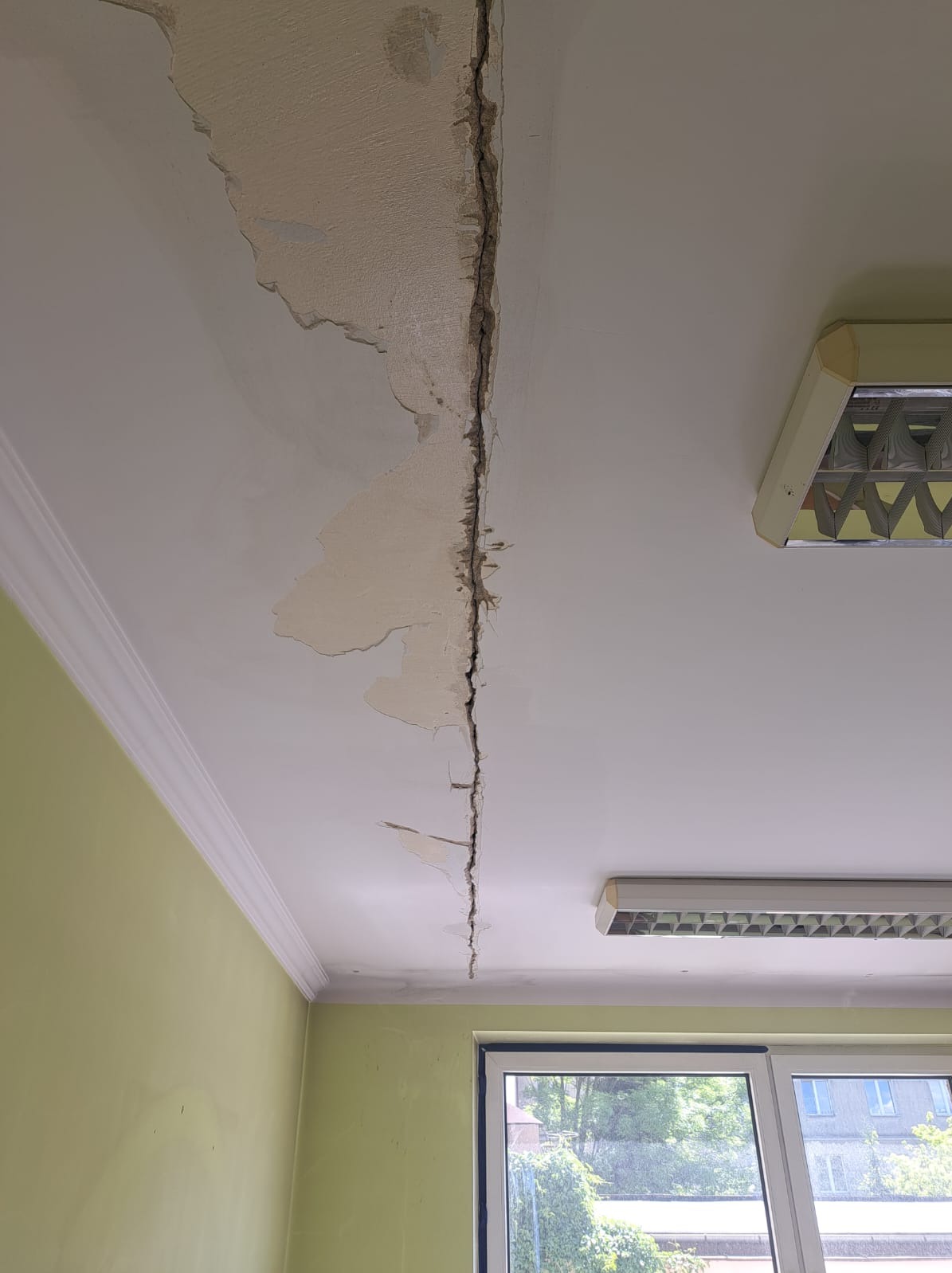 Pęknięcie i odpadający tynk na suficie w pomieszczeniu z oknem i lampą fluorescencyjną, widoczne uszkodzenia wymagające naprawy.