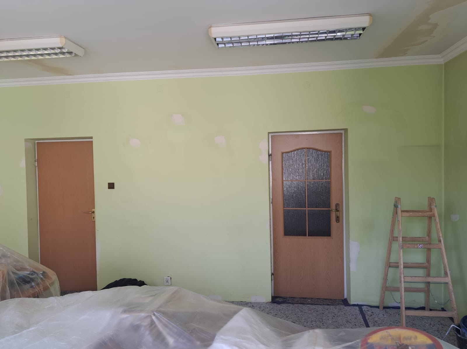 Pokój w trakcie szpachlowania ścian, z widocznymi łatami szpachli, zabezpieczoną folią podłogą, drewnianą drabiną i dwoma drzwiami.