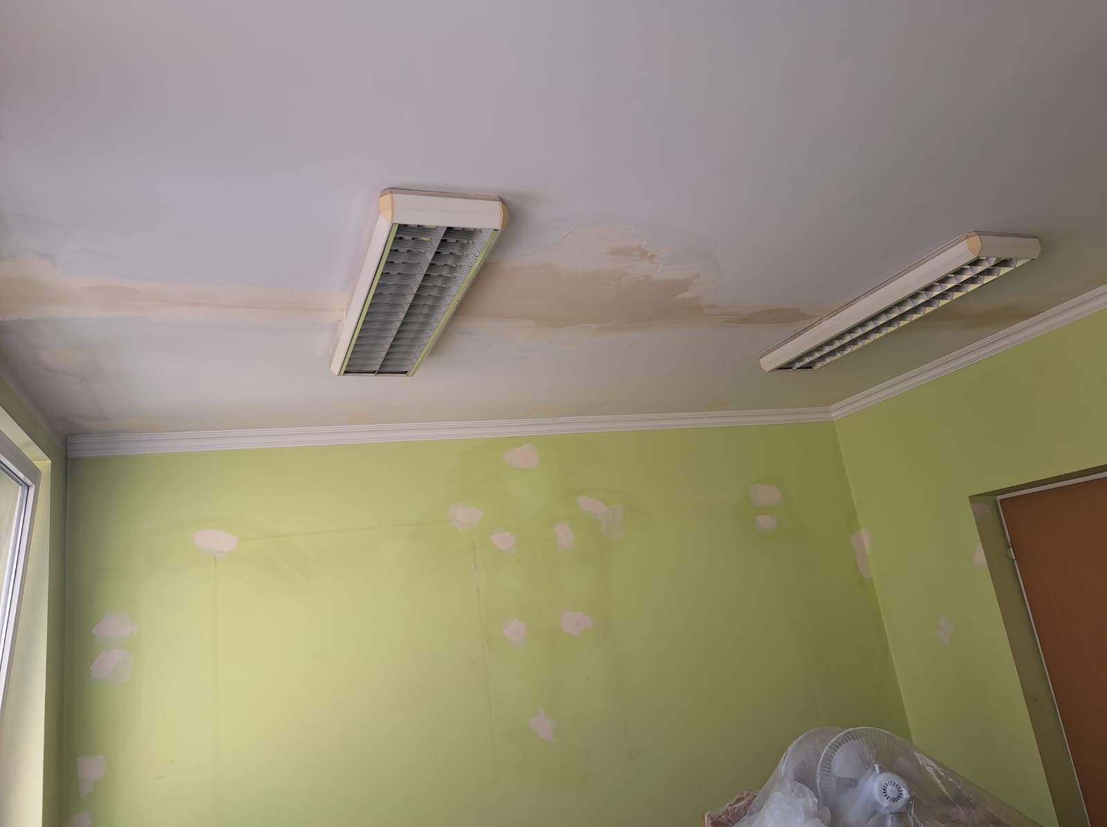 Pokój z zielonymi ścianami w trakcie przygotowania do malowania, widoczne plamy po szpachlowaniu i zacieki na suficie, oświetlenie jarzeniowe.