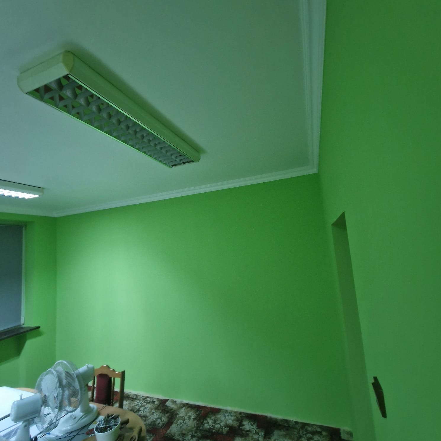 Świeżo pomalowane na jaskrawy zielony kolor ściany pokoju z widocznym oknem, lampą sufitową i starym wentylatorem.