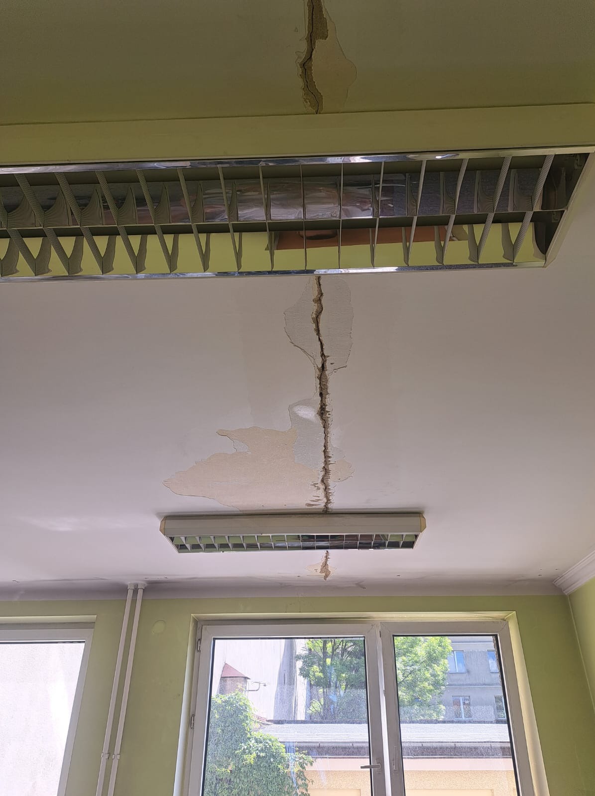 Pęknięty sufit w pomieszczeniu z oknem, widoczne ślady po zalaniu i odpadający tynk, oświetlenie rastrowe.