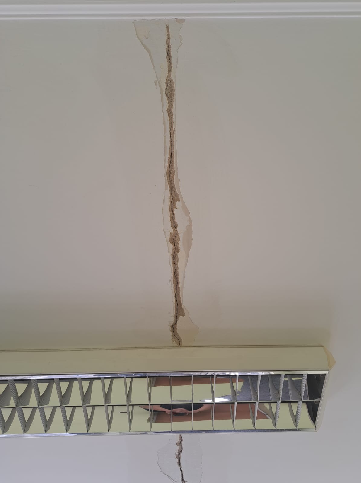 Pęknięcie na suficie z widocznymi warstwami uszkodzonego materiału, przebiegające od listwy sufitowej do lampy, z mniejszym pęknięciem poniżej lampy.