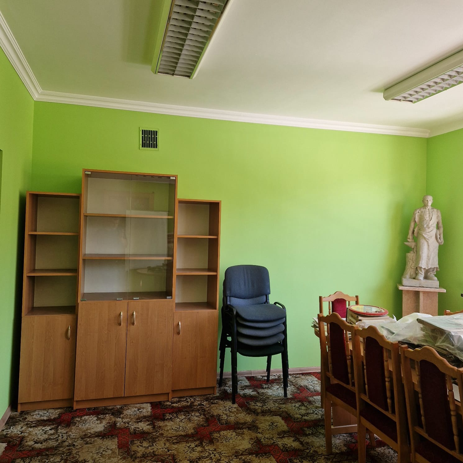 Wnętrze biura z zielonymi ścianami, drewnianymi regałami, krzesłami w stosie i rzeźbą na postumencie, dywan z wzorem i oświetlenie jarzeniowe.