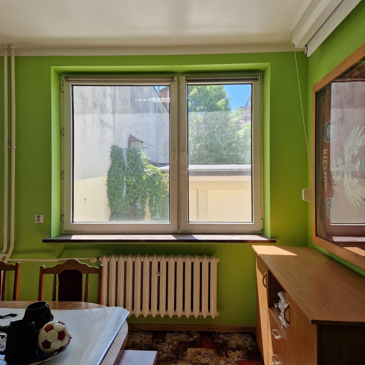 Jasnozielony pokój z oknem wychodzącym na budynek porośnięty bluszczem, grzejnik pod oknem, drewniany kredens i stół z piłką do nogi.