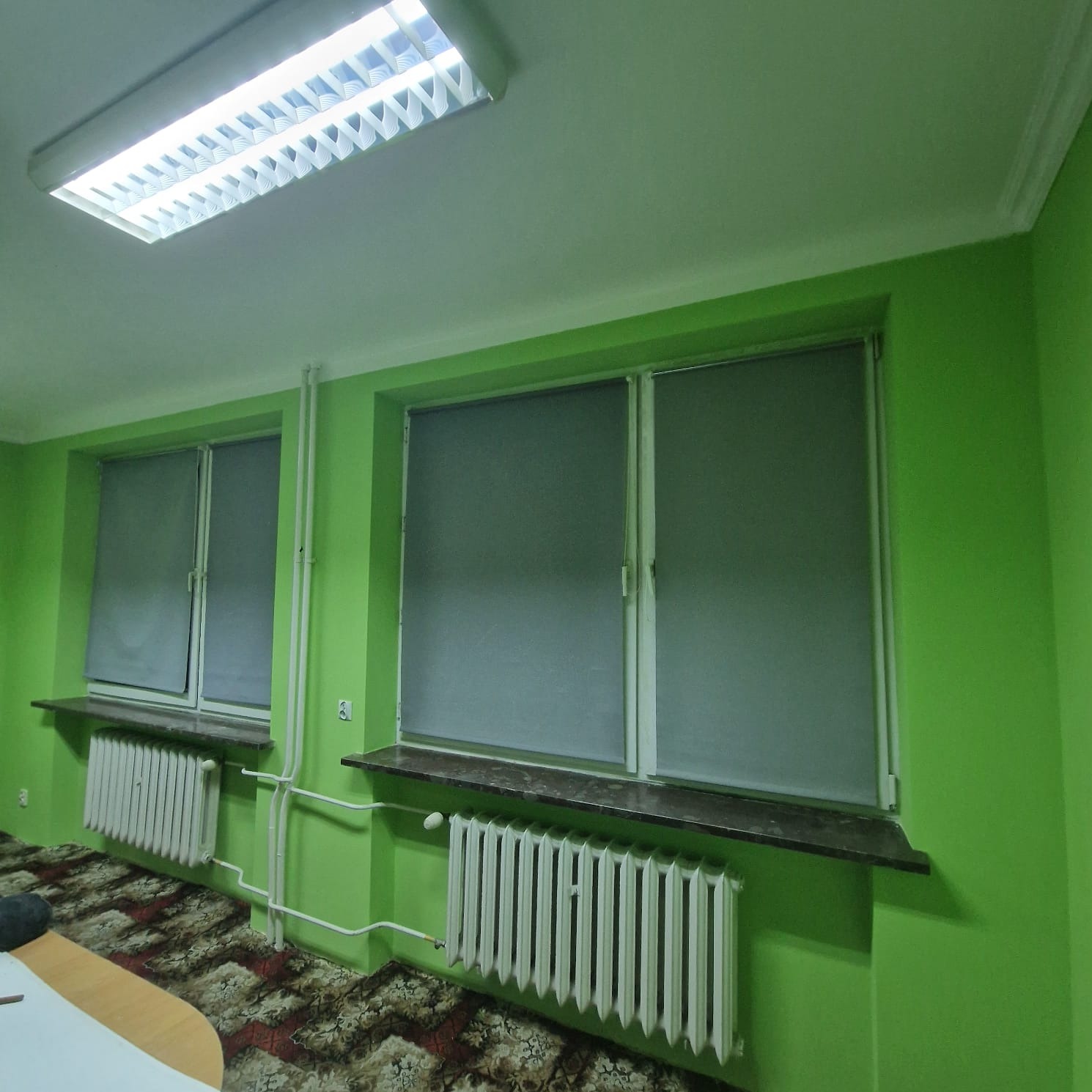 Jasnozielone wnętrze pokoju po odświeżeniu, z dwoma oknami z roletami, białymi grzejnikami pod parapetami i wzorzystą podłogą.