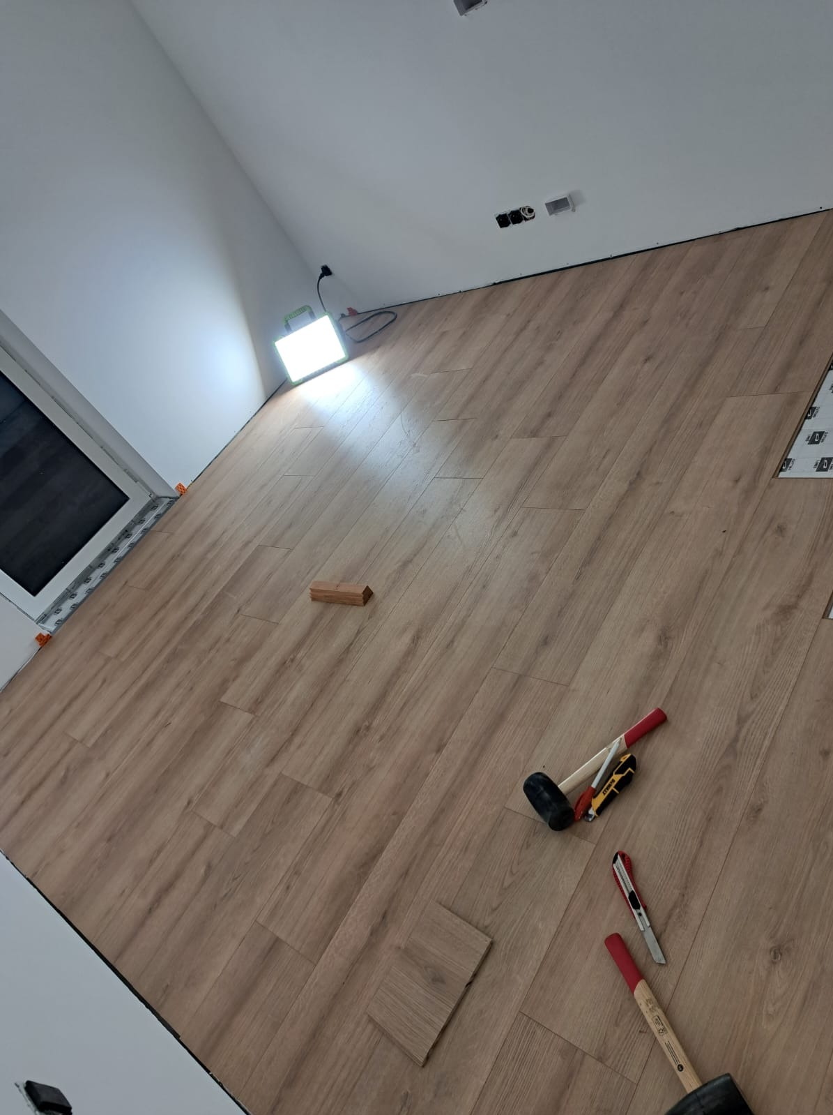 Układanie paneli podłogowych w pomieszczeniu, widoczne narzędzia: młotek gumowy, nożyki, kliny dystansowe, oświetlenie halogenowe.