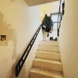 Wnętrze klatki schodowej w trakcie remontu z zamontowaną czarną poręczą metalową na ścianie, widoczne niedoskonałości ścian i schodów.