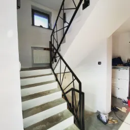 Surowe schody z pomalowanymi na biało betonowymi stopniami i czarną, geometryczną balustradą metalową, widoczne niedoskonałości wykończenia.