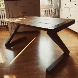 Stół z drewnianym blatem i metalowymi nogami w kształcie litery Z, stojący na drewnianej podłodze w jasnym wnętrzu.