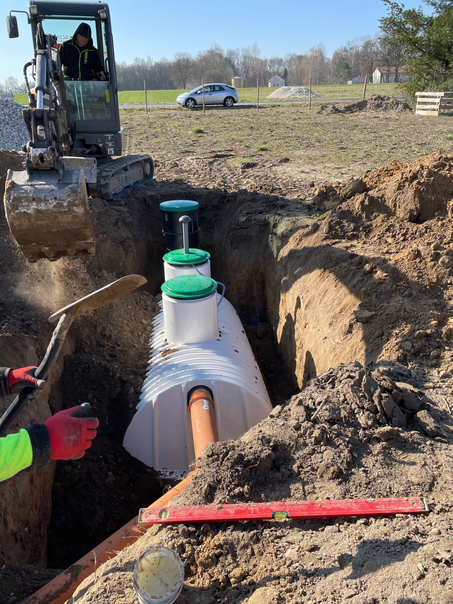 Montaż przydomowej oczyszczalni ścieków: biały zbiornik z zielonymi pokrywami umieszczony w wykopie, widoczny poziomica, rury kanalizacyjne, łopata i pracujący operator koparki w tle.