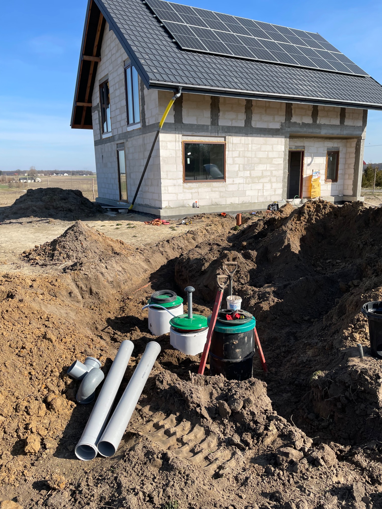 Montaż przydomowej oczyszczalni ścieków na działce z surowym budynkiem, widoczne białe zbiorniki z zielonymi pokrywami, rury kanalizacyjne i wykop ziemny.