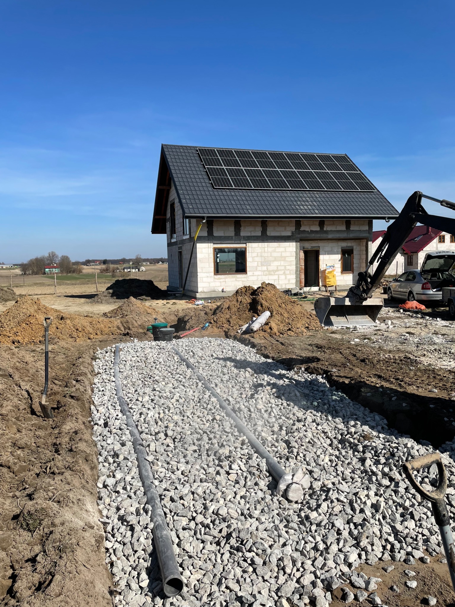 Instalacja drenażu rozsączającego: rury drenarskie ułożone w rowie wypełnionym grysem na tle powstającego domu z panelami słonecznymi na dachu i koparki.