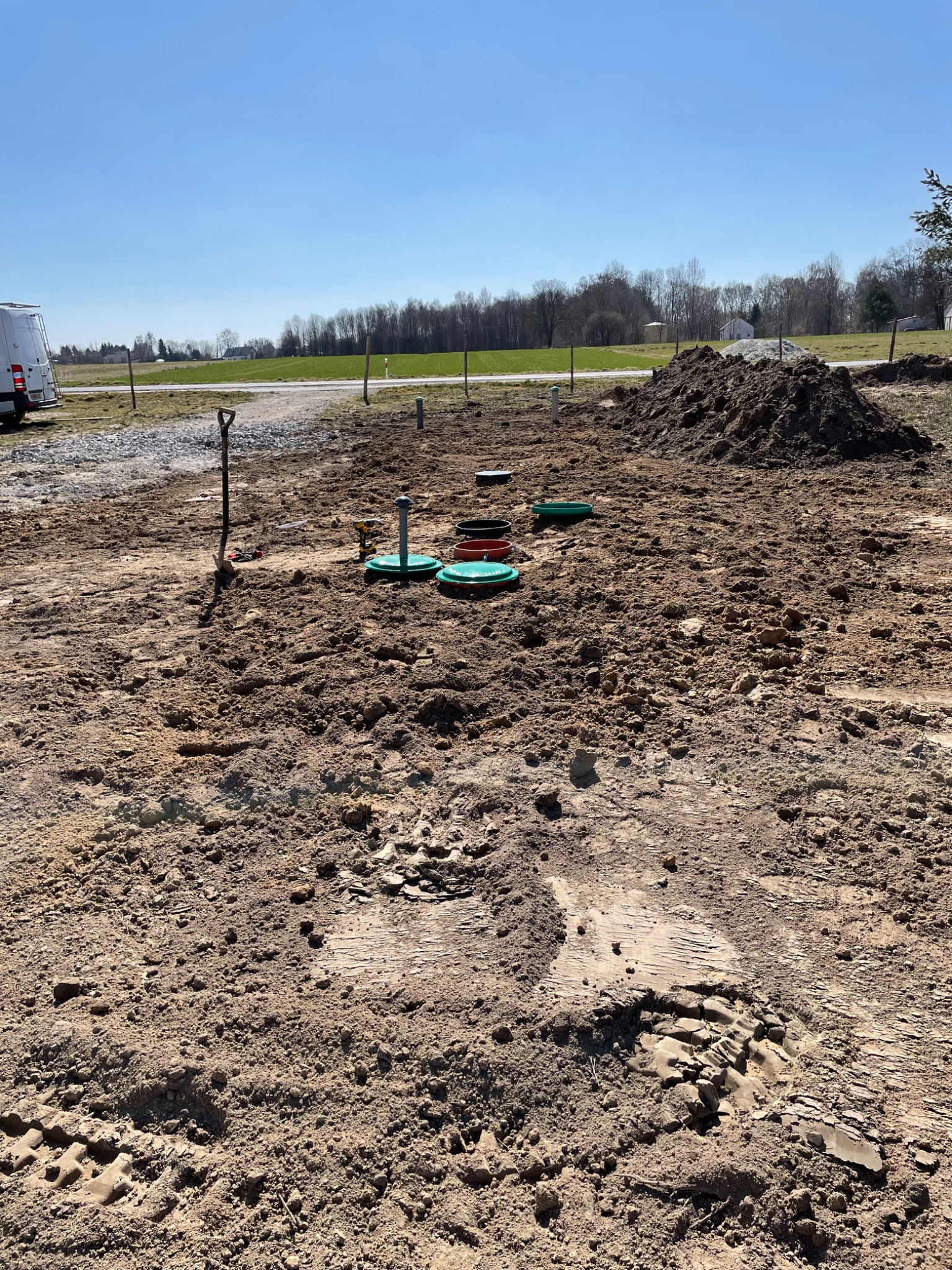 Teren po wykonanych pracach ziemnych, widoczne włazy do instalacji podziemnych w kolorach zielonym i czerwonym, oraz narzędzia budowlane i sterta ziemi.