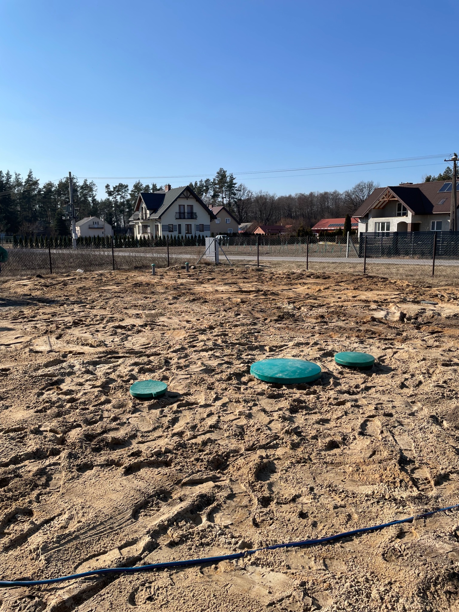 Teren po budowie przydomowej oczyszczalni ścieków z widocznymi zielonymi pokrywami studzienek i śladami opon na piasku, w tle zabudowa jednorodzinna i drzewa.