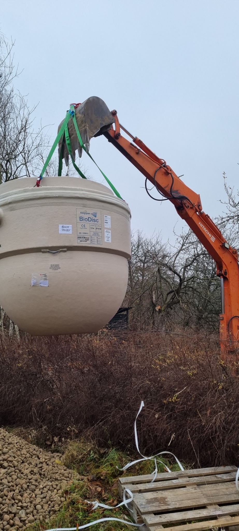 Pomarańczowa koparka Hitachi podnosi beżowy zbiornik BioDisc na tle jesiennego krajobrazu z brązowymi krzewami i nagimi drzewami.