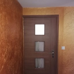 Dar-but firma handlowo usługowa Dariusz Czajkowski - Brązowe drzwi z trzema kwadratowymi okienkami na tle ściany pokrytej dekoracyjną, złoto-brązową fakturą przypominającą pomarszczony papier.